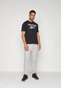 Must Reeboki T-särk valge logoga, kombineeritud heledatehallide dressipükstega. Mustad spordijalatsid lõpetavad outfiti.