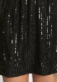 Svart klänning med vertikala veck, prydd med glittrande paljetter som skapar en texturerad yta. Sömmen är scallopad, vilket ger en elegant look.