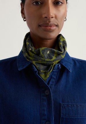 Zelený květinový šátek z měkké látky, obtočený kolem krku, v kombinaci s modrou džínovou košilí s knoflíkovou přední částí a kapsou.