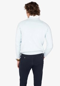 Hombre con cabello rizado que lleva un suéter de color azul claro y pantalones oscuros, mostrado de espaldas contra un fondo blanco liso.