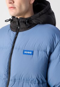 Light blue puffer jacket with a black hood, featuring a front zipper and a HUGO logo patch in blue. Textured fabric with horizontal seams.