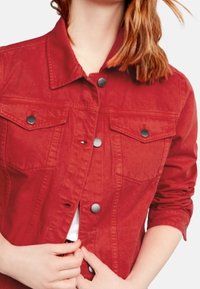 Rød denimjakke med en tætsiddende pasform, prydet med metaltryklåse, to brystlommer og krave. Overfladen ser glat ud.