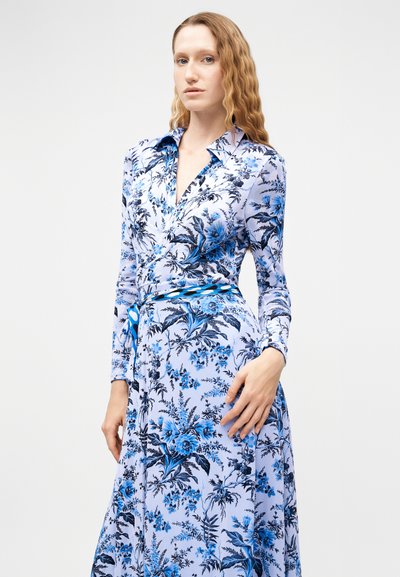 Γυναίκα με κυματιστά μαλλιά που φοράει μακρυμάνικο, μπλε και λευκό φόρεμα τύπου wrap με λουλουδάτο μοτίβο, γιακά και ζώνη στη μέση.