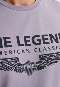 Fialové bavlněné tričko s odvážným černým textem "LEGEND" a "AMERICAN CLASSIC." Grafika křídel a štítu dodává na detailu.
