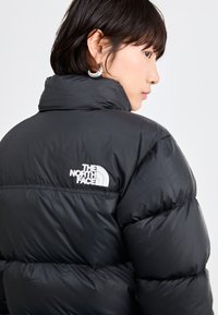 Musta puffatakki korkealla kauluksella. Sisältää valkoisen The North Face -logon takana, teksturoituja ja pehmustettuja osia eristykseen.