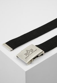 Svart vävd tygbälte med en silvermetallspänne som har den gravyr som säger "ALPHA INDUSTRIES USA" och en geometrisk design.