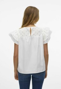 Donna di spalle che indossa una camicetta bianca con yoke in pizzo occhio di pernice e maniche a volant, abbinata a jeans blu, su sfondo semplice.
