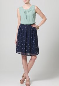 Blusa senza maniche verde menta con pieghe, abbinata a una gonna blu scuro con motivo di uccelli bianchi. Sandali beige con cinturini completano l'outfit.