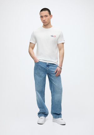 Jeune homme aux cheveux courts portant un t-shirt blanc à motif, un jean large bleu clair, des baskets blanches et un bracelet en argent, debout les mains dans les poches.