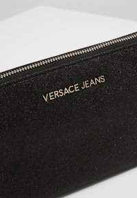 Pochette noire en matière pailletée texturée, équipée d'une fermeture éclair ton or et d'un logo embossé "VERSACE JEANS" sur le côté.