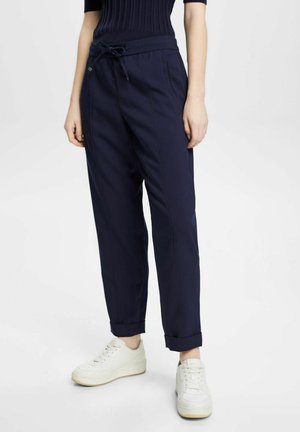 Pantalones azul marino de tejido suave, con cintura ajustable, bolsillos laterales y puños remangados, combinados con zapatillas blancas.