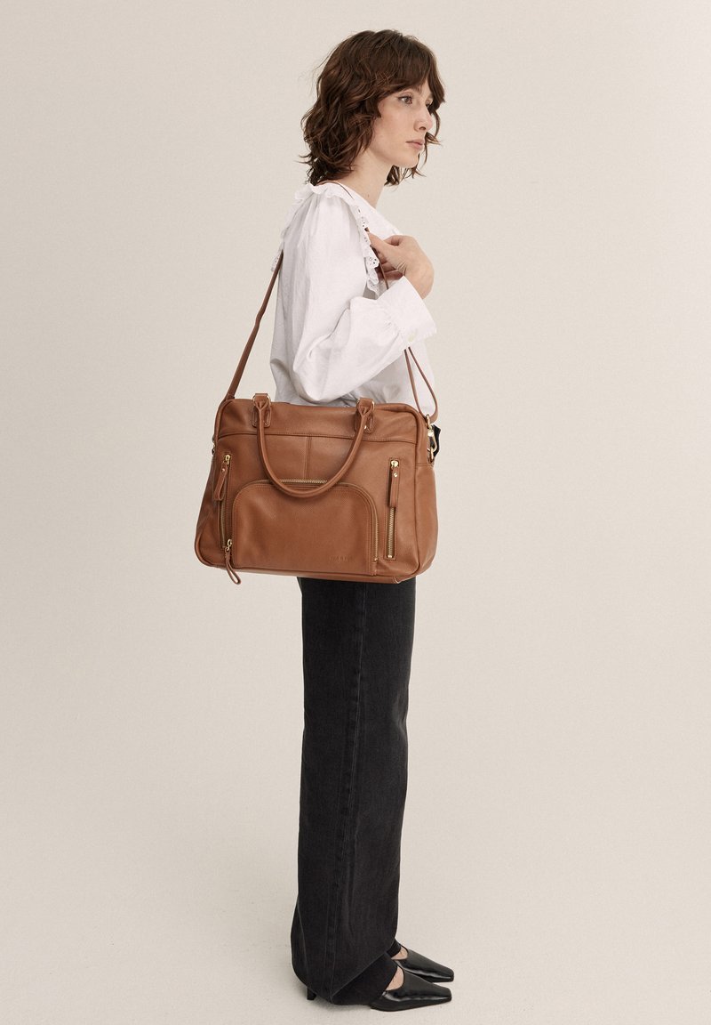 Borsa tote in pelle marrone con manici doppi e accessori in tono oro. Presenta una tasca frontale con zip e una superficie liscia. Modello che indossa jeans neri.