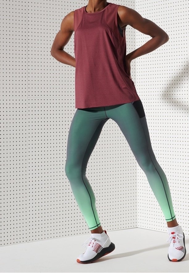 Sportowy top na ramiączkach w kolorze bordo, zestawiony z legginsami w zielonej gradacji oraz białymi butami sportowymi z czerwonymi akcentami. Gładki materiał, dopasowany krój.