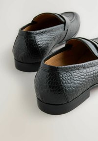 Schwarze Lederloafer mit einem strukturierten, reptilienartigen Muster, einer flachen Gummisohle und einem gesteppten Akzent entlang der oberen Kante.