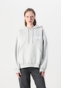 Lyse grå hettegenser med frontlomme, snorhetten og ribbestrikkede mansjetter. Har et minimalistisk hvitt grafisk design på brystet. Brukt over mørke jeans.