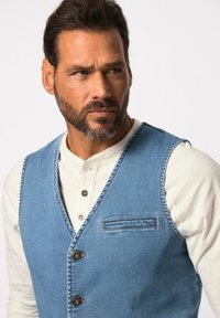 Gilet in denim di un blu chiaro con scollo a V profondo, quattro bottoni e una tasca sul petto. Indossato sopra una maglietta henley bianca.