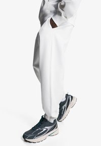 Personne portant un pantalon de survêtement blanc et des chaussures de sport noir et blanc, se tenant avec les mains dans les poches devant un fond blanc.