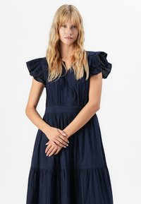 Mladá žena s dlouhými blond vlasy, oblečená v tmavě modrých šatech s volnými rukávy, stojící s jemně sepnutýma rukama vpředu.
