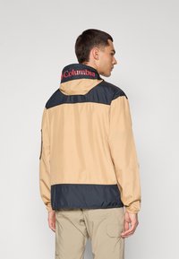 Beige og marineblå vindjakke med hette, designet med høy krage, elastiske mansjetter og logo på nakken. Glatt, lett materiale.