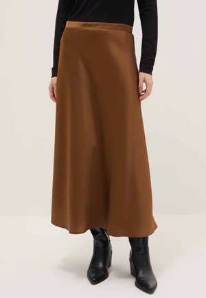 Frau trägt einen langen braunen Rock, ein schwarzes langärmliges Oberteil und schwarze Stiefel mit Absatz, steht vor einem schlichten hellen Hintergrund.