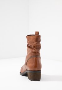 Braune Lederstiefel bis zur Wade mit gestapeltem Blockabsatz, Zugschlaufe oben und leicht zerknittertem Schaft, von hinten auf einer weißen Oberfläche betrachtet.