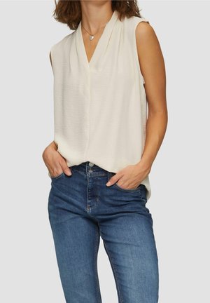 Žena v krémovém halence bez rukávů zastrčené do modrých džínů s vysokým pasem, ruce v kapsách, před neutrálním pozadím.