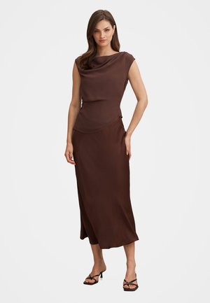 Vrouw staand, draagt een mouwloze bruine satijnen top met gedrapeerde halslijn en bijpassende lange rok, gecombineerd met zwarte sandalen met bandjes.