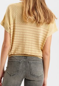 Krótka bluzka z krótkim rękawem w złote paski z teksturowanym wzorem dzianiny; do tego szare dżinsy z dwoma tylnymi kieszeniami.
