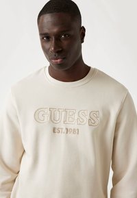 Sudadera de cuello redondo en beige claro con el logotipo bordado "GUESS EST. 1981" en hilo tono sobre tono. Tejido suave con una textura sutil.