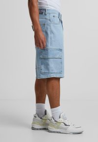 Lyseblå denim cargo-shorts med to store sidelommer og et merke-logo på linningen, kombinert med hvite joggesko og hvite sokker.