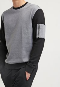 Sweat-shirt gris et noir avec un col rond, présentant une texture côtelée et une poche zippée sur la manche gauche.
