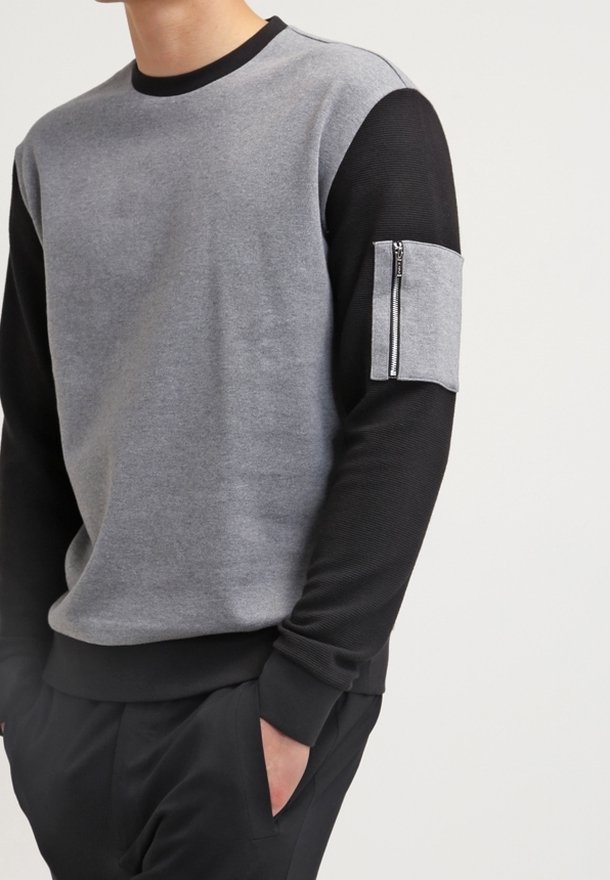 Sweat-shirt gris et noir avec un col rond, présentant une texture côtelée et une poche zippée sur la manche gauche.