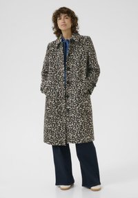Manteau en imprimé léopard brun et beige, avec un col, des poches avant et des boutons sur le devant. Associé à un pantalon en denim foncé.