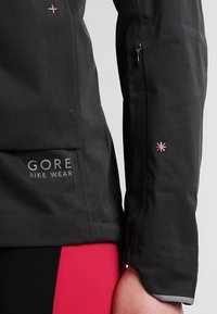 Manga de chaqueta de ciclismo negra mostrando el logo "GORE BIKE WEAR", detalle de estrella bordado en rosa y pantalones de ciclismo en rojo y negro.