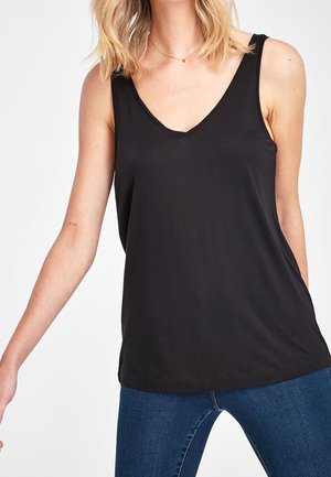 Femme aux cheveux blonds mi-longs portant un haut noir sans manches à col en V et un jean bleu sur un fond uni.