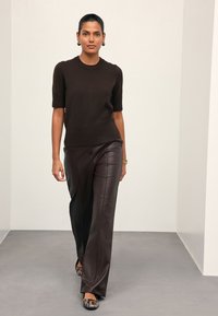 Femme marchant avec un pull noir à manches courtes, un pantalon en cuir marron foncé, une montre en or et des chaussures à motif peau de serpent sur fond uni.