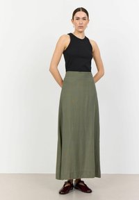 Top senza maniche nero abbinato a una gonna verde oliva a vita alta e lunga fino al pavimento. La modella indossa scarpe bordeaux, in piedi davanti a uno sfondo neutro.