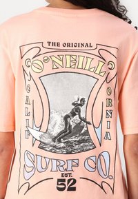 Jasnoróżowa koszulka bawełniana z graficznym wzorem przedstawiającym surfingu. Tekst zawiera "O'Neill Surf Co." oraz "The Original California" w stylu retro.
