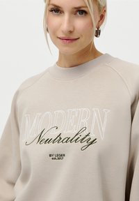Blond kvinne som har på seg en beige hettegenser med hvit og grønn tekst "MODERN Neutrality BY LEGER est. 2017" og sølv hoop-øreringer.