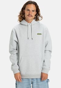 Sudadera gris claro de tela suave, con un bolsillo tipo canguro, cordones y un pequeño parche de logo verde en el pecho.