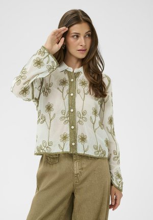 Žena v priesvitnej bielej blúzke s olivovo zelenou kvetinovou výšivkou a vysokými olivovo zelenými nohavicami, pózujúca s rukou pri čele.