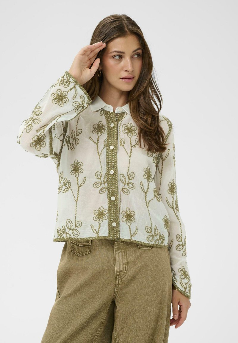 Sieviete valkā caurspīdīgu baltu blūzi ar olīvu zaļas krāsas ziedu izšuvumiem un augsta vidukļa olīvu zaļas krāsas bikses, pozējot ar roku pie pieres.