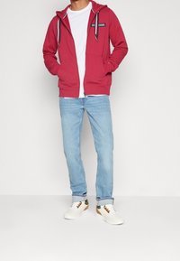 Sweatshirt rouge à capuche avec poches avant, fermeture éclair blanche et accents bleu marine. Associé à un jean bleu clair et des baskets blanches avec des accents marron.