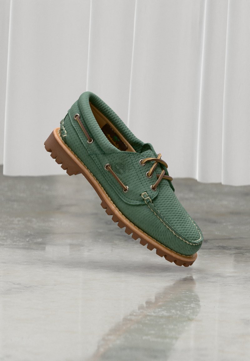 Chaussure bateau en cuir texturé vert avec lacets marron et semelle épaisse en caoutchouc marron, posée sur un sol clair avec un fond de rideau blanc.