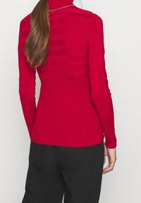 Pull à col roulé rouge avec texture côtelée et accents de rayures horizontales dans le dos, associé à un pantalon noir.