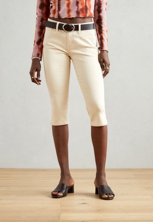 Jeans cortos beige con una textura sutil, cinco bolsillos y un corte clásico, combinados con sandalias de tacón bloque abiertas en la parte delantera de color negro.