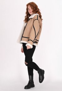 Beige takki, jossa on lampaanvillavuori, mustat yksityiskohdat, ylisuuri malli. Yhdistetty revenneisiin mustiin farkkuihin ja tukevien mustien saappaiden kanssa.