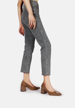 Jeans grigi alti in vita con orlo grezzo, abbinati a scarpe con tacco a blocco in suede marrone, caratterizzate da una punta arrotondata e un accento metallico.