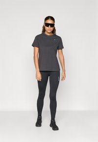 Szürke póló kék logóval, fekete leggings-szel és fekete sportcipővel párosítva, modell fekete napszemüvegben, egyszínű háttér.