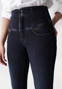 Detailní záběr na tmavě modré džíny s vysokým pasem a dvěma knoflíky na zapínání, s bočními kapsami, nošeno s bílou košilí s ohrnutými rukávy.